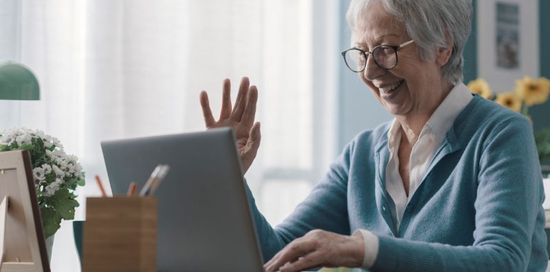 Ältere Dame sitzt vor Ihrem Computer und winkt in die Kamera.